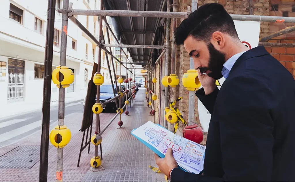  Controle de qualidade na construção civil: A imagem mostra um homem com barba vestindo um traje e gravata, sentado em frente a um prédio em construção, lendo um papel na mão. Ele está usando um capacete e óculos de proteção. Existem tapumes e escadas em segundo plano. 