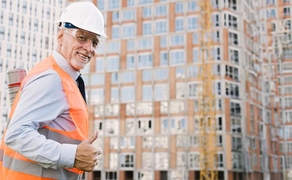 Controle de qualidade na construção civil: A imagem mostra um homem com uma camisa de proteção e um capacete de segurança em pé diante de um local de construção com edifícios altos no fundo. Ele está dando um sinal de que tudo está bem, indicando que tudo está em ordem. A imagem é tirada de uma angulação baixa, dando a impressão de um grande local de construção com edifícios altos no fundo. O homem está vestindo uma camisa de proteção e um capacete de segurança, indicando que ele é um trabalhador de construção. A imagem é tomada durante o dia, com o sol brilhando forte no fundo. A imagem é clara e bem iluminada, sem visíveis imperfeições ou distorções.