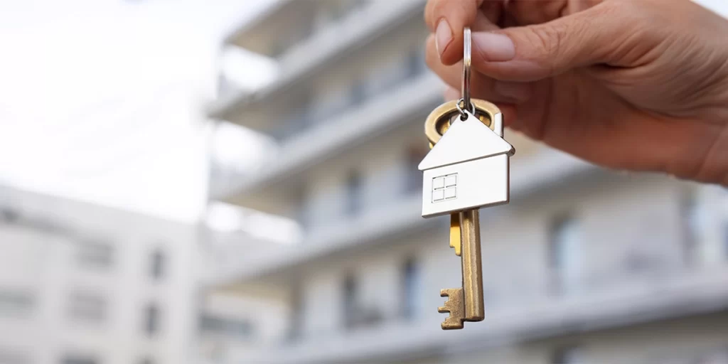 Open House: A imagem mostra uma mão humana segurando uma chave de corrente. A chave tem um acabamento com um logotipo de uma casa, sugerindo que pode ser uma chave de uma casa. A chave é de cor brilhante, possivelmente metal, e está pendurada por um cabo que parece ser um corrente. A mão está na parte superior da chave, e a chave está na parte inferior da mão, indicando que a chave está sendo segurada ou mostrada. Ao fundo, há uma estrutura que parece ser um edifício, mas a foco da imagem é na chave e na mão, o que faz com que o fundo esteja desfocado e não seja possível identificar detalhes específicos do edifício.