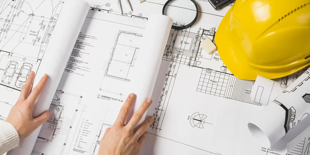 as built: A imagem mostra duas mãos de uma pessoa abrindo e examinando um livro de projetos arquitetônicos. Ao fundo, há outros documentos de projeto e equipamentos relacionados ao setor de construção, como um capacete amarrado e um estetoscópio. A cena sugere um ambiente de trabalho de projeto de construção ou arquitetura.