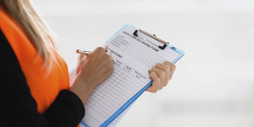 Checklist de obra: A imagem mostra uma pessoa de perfil preenchendo um formulário de registro. A pessoa está vestindo uma camiseta laranja e parece estar focada em preencher o formulário. O formulário é um caderno azul com uma folha de papel preenchida com informações, e a pessoa está usando uma caneta para escrever. A cena sugere uma situação de registro ou inscrição, possivelmente em um evento ou em uma instituição. A foto foi tirada de cima, focando na mão da pessoa e no formulário, com um fundo desfocado que não oferece informações adicionais.