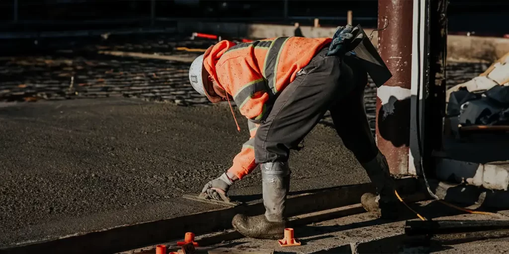 concreto usinado: A imagem mostra uma pessoa em um ambiente de construção. A pessoa está de costas para a câmera, com uma postura baixa, sugerindo que está trabalhando. Ela está vestindo um traje de segurança que inclui uma jaqueta amarronada e um capacete, indicando que está em um ambiente de trabalho onde a proteção é necessária. A jaqueta tem detalhes de segurança, como linhas amarelas e vermelhas, que são comuns em trajes de segurança para aumentar a visibilidade. A pessoa está usando calças pretas e parece estar usando algum tipo de protetor para as mãos, possivelmente para segurança contra ferramentas ou materiais.