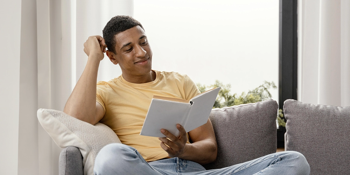 conforto ambiental: A imagem mostra um jovem sentado em um sofá, lendo um livro. Ele está usando uma camiseta amarela e calça jeans azul claro. O jovem tem uma expressão séria e concentrada, com uma mão sobre o queixo e o outro segundo o livro. Ele está de pé no sofá, com as pernas cruzadas. Atrás dele, há uma janela com cortinas abertas, permitindo que luz natural entre, e um ambiente aparentemente tranquilo e acolhedor.