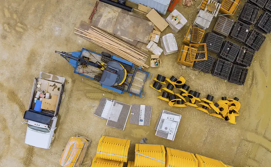 logística de canteiro de obras: Esta imagem mostra uma pilha de materiais de construção, incluindo uma grande pilha de beams de madeira, uma pilha de blocos de cimento e uma pilha de pedras. Existem também um grande excavador no primeiro plano e um pequeno caminhão parado próximo. A imagem é tirada de uma altura elevada, olhando para baixo no local de construção. O fundo é composto por terra e lixo, e há algumas árvores no horizonte. A cena geral é caótica e desorganizada, com muitos materiais e equipamentos espalhados por lá.