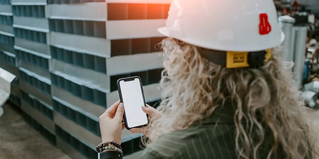 materiais inovadores: A imagem mostra uma pessoa de cabelo longo e ondulado, vestindo um capacete branco com um logotipo vermelho no lado direito. A pessoa está segurando um smartphone na mão direita, e o ecrã do dispositivo está iluminado, mas não é possível ver o conteúdo exibido. A pessoa está de pé, e ao fundo, há uma estrutura que parece ser parte de uma construção ou obra, com ladrilhos ou blocos de construção em tons de azul e cinza. A luz do sol está refletindo na parte superior da imagem, sugerindo que a foto foi tirada durante o dia.