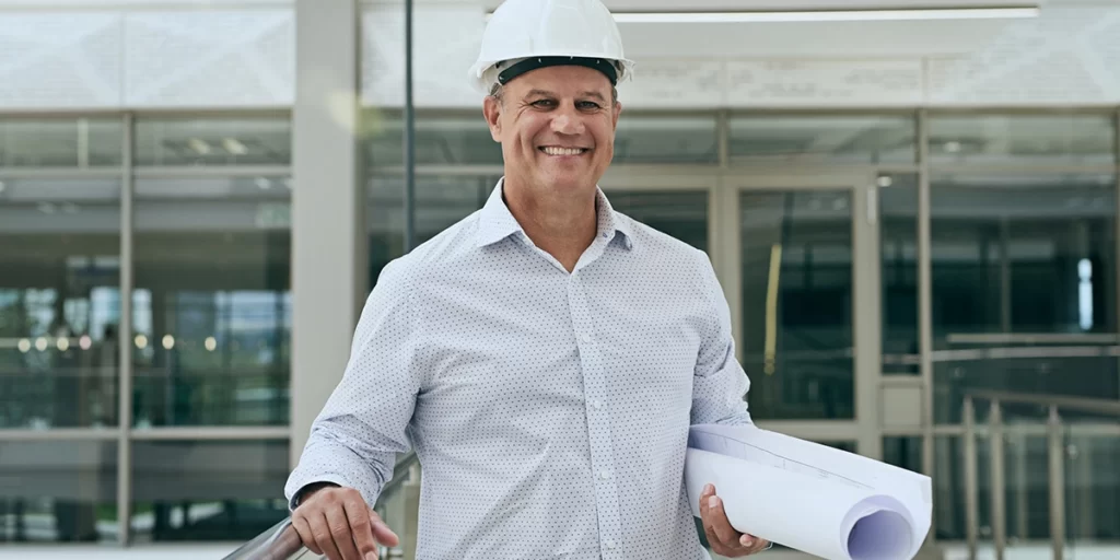 software de gestão para construtoras: A imagem mostra um homem usando um capacete branco e segurando um conjunto de planilhas. Ele está em uma escada levando para um prédio com grandes janelas e um design moderno. O homem está sorrindo e parece feliz. O fundo é um prédio moderno com janelas de vidro e uma fachada branca. A imagem está bem iluminada e as cores são vibrantes e vivas. A atmosfera geral da imagem é alegre e otimista.