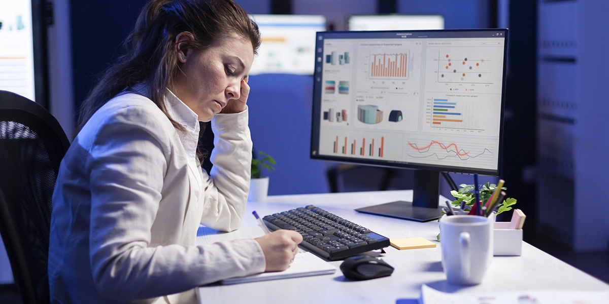 software de gestão para construtoras: A imagem mostra uma mulher sentada em frente a dois monitores de computador. Ela está usando uma camisa branca e pantalones negros, e tem a cabeça em seus braços. Há várias gráficos e tabelas exibidas nos monitores em frente dela. A sala está pouco iluminada, com algumas luzes em cima do escritório e dos monitores. A mulher parece cansada e estressada, com uma expressão preocupada no rosto. A atmosfera geral da imagem é de tensão e estresse.