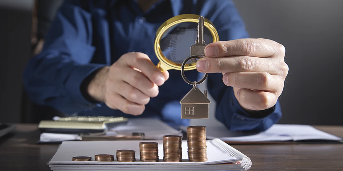 Como vender Imóveis para investidores: imagem das mãos de uma pessoa segurando uma lupa com uma mão e um chaveiro de casa na outra. À frente, numa mesa vemos pilhas de moedas em ordem crescente.