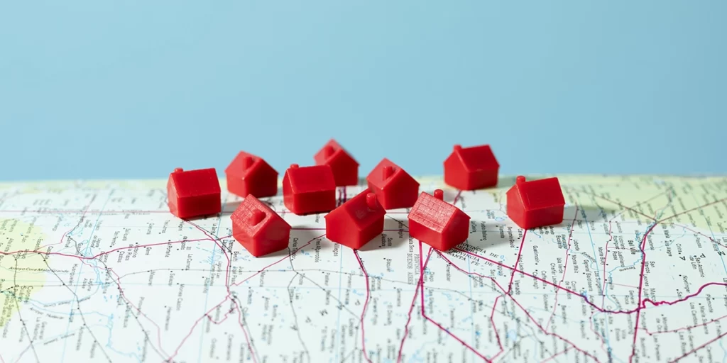 Gerenciamento de Leads: A imagem mostra uma série de pequenas estruturas que parecem ser modelos de casas ou edifícios, todos iguais e de cor rosa. Eles estão dispostos sobre um mapa físico, que parece ser um mapa de um país ou região, com linhas que representam caminhos ou vias. O fundo é azul, sugerindo que o mapa está sobre um suporte ou um papel de cor azul. A imagem tem um estilo simples e é focada na representação dos modelos de casas sobre o mapa, possivelmente para ilustrar uma ideia de urbanização ou planificação urbana.