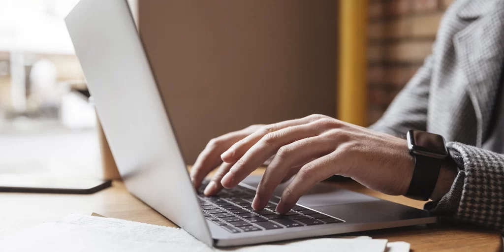 Marketing de conteúdo para construtoras: A imagem mostra uma pessoa sentada em frente a um laptop aberto. A pessoa está usando um teclado externo. Ela está vestindo uma camisa com um padrão que parece ser um terno ou uma jaqueta de tecido, o que pode indicar um ambiente profissional ou acadêmico. A mão da pessoa está sobre o teclado, e ela parece estar digitando ou segurando o teclado. Há uma página de papel aberta ao lado do laptop, mas não é possível ler o conteúdo. A foto foi tirada de cima, focando na mão e no teclado, e a cena está desfocada para enfatizar a atividade da mão. A luz natural parece estar iluminando a cena, sugerindo que a foto foi tirada durante o dia.