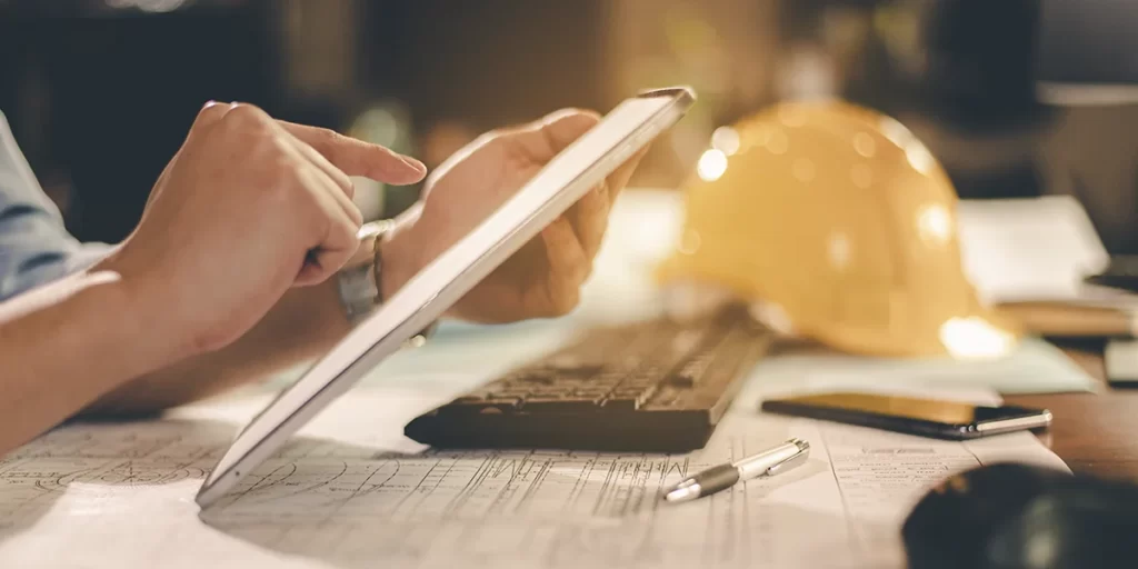 cronograma fisico e financeiro de obras: A imagem mostra uma mão humana ao lado de um tablet de tela grande, que está aberto e colocado sobre um teclado. A mão está segurando o tablet, e parece estar a tocar ou interagir com o ecrã. Há um ambiente de escritório ao fundo, com uma luz suave que ilumina a parte superior do tablet e a mão, criando um efeito de profundidade de campo. Há também um capacete de construção ao fundo, sugerindo que a pessoa pode estar trabalhando em um projeto de construção ou estar em um ambiente de trabalho relacionado ao setor de construção. A imagem tem um filtro que dá uma sensação de calma e concentração.