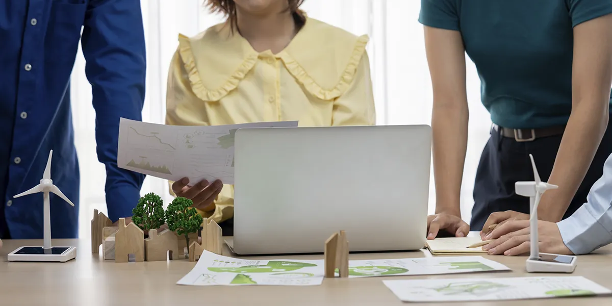 o que torna uma construção sustentável: imagem de três pessoas diante de uma mesa de escritório. O ângulo da câmera foca nos itens em cima da mesa.