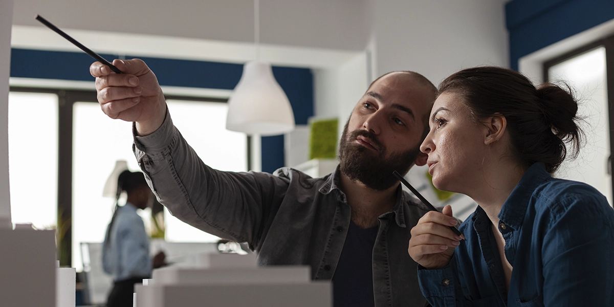 A imagem apresenta um homem e uma mulher sentados um ao lado do outro, ambos com um lápis na mão. O homem está direcionando o lápis para frente e os dois olham para a mesma direção.